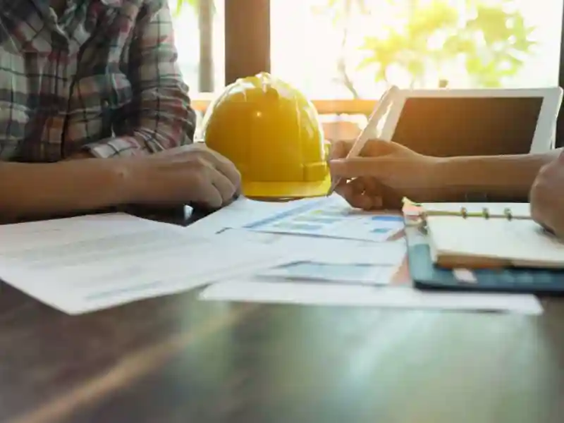 Two individuals discussing documents on a table with a yellow hard hat, papers, and a tablet.